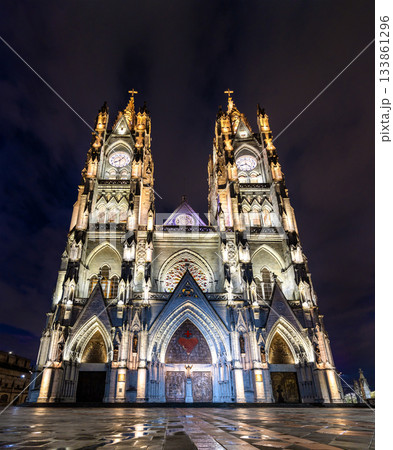 Illuminated facade of Basilica del Voto Nacional rises above wet plaza in Quito, Ecuador. Neo-Gothic church features twin towers and intricate stone carvings under dark night sky 133861296