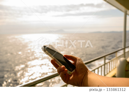 朝日を浴びるフェリーの船上でスマートフォンを持つ男性の手元（船名：おき） 133861312