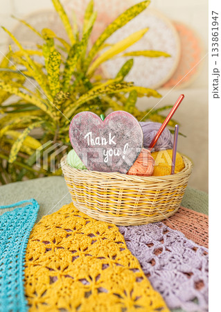 Basket of colorful yarn and crochet hooks and red wooden heart with Thank you lettering on background of surrounded colorful crocheted fabric and home flowers Basket of colorful yarn and crochet hooks and red wooden heart with Thank you lettering on background of surrounded colorful crocheted fabric and home flowers 133861947