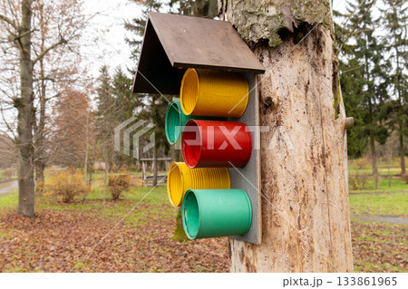 A bird feeder made of colorful canisters is fixed to a tree in a park. Leaves have fallen, and the ground is covered in autumn foliage 133861965