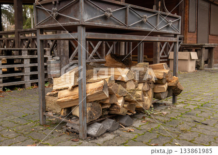A collection of neatly stacked firewood rests on a metal rack outside a wooden structure, surrounded by fallen leaves and stones 133861985