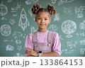 A young kid with arms crossed smiles in front of a chalkboard with drawings of rockets and planets. 133864153