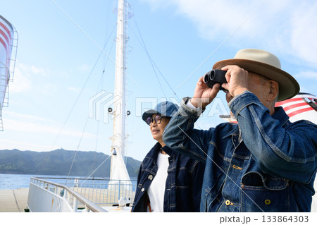 双眼鏡で景色を眺めるデニム姿のシニア夫婦、フェリーのデッキで船旅を楽しむ 133864303