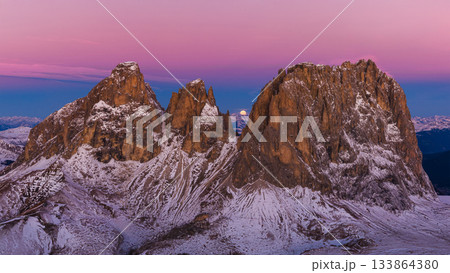 Panoramic view of Dolomite mountain peaks under a glowing full moon. Early winter landscape with snow-covered summits in soft dawn light. Panoramic view of Dolomite mountain peaks under a glowing full moon. Early winter landscape with snow-covered summits in soft dawn light. 133864380