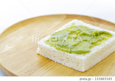 A single slice of white bread with a spread of Pandan Custard is placed on a round wooden plate, showcasing simple and natural A single slice of white bread with a spread of Pandan Custard is placed on a round wooden plate, showcasing simple and natural 133865585