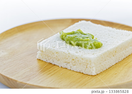 A close-up of a thick slice of white bread with a Pandan Custard placed on top, presented on a round wooden plate against a clean A close-up of a thick slice of white bread with a Pandan Custard placed on top, presented on a round wooden plate against a clean 133865828