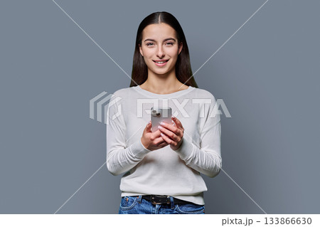 Portrait of smiling teenage girl holding smartphone on gray studio background Portrait of smiling teenage girl holding smartphone on gray studio background 133866630