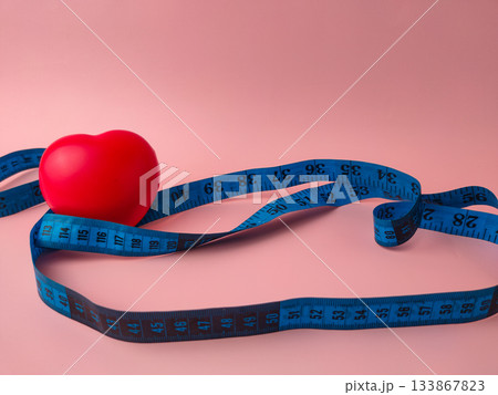 The image features a red heart object encircled by a blue tape measure, set against a pink background. The image features a red heart object encircled by a blue tape measure, set against a pink background. 133867823