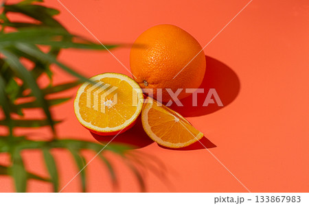 On the coral surface, next to two neatly sliced slices, there is a whole orange. Sunlight casts soft shadows, emphasizing the freshness and bright color of the fruit. 133867983