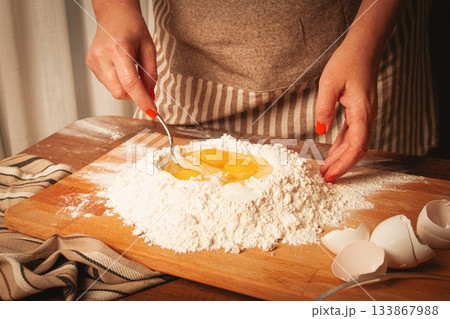 Mixing eggs into flour to make dough for homemade pasta in a cozy kitchen setting 133867988