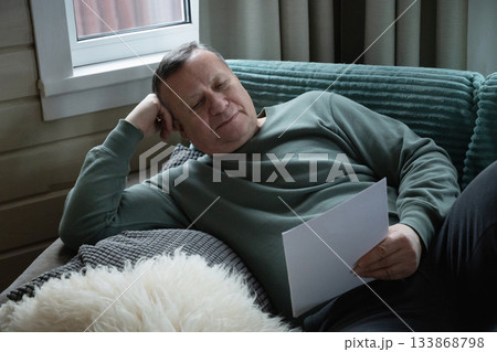 Resting caucasian man lying on sofa and reading paper, enjoying a quiet moment and relaxation indoors in a cozy home space. Home Leisure Activities 133868798