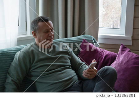 Relaxing caucasian man sitting on sofa and using smartphone indoors, enjoying a calm moment in a cozy home environment. Home Leisure Activities 133868801
