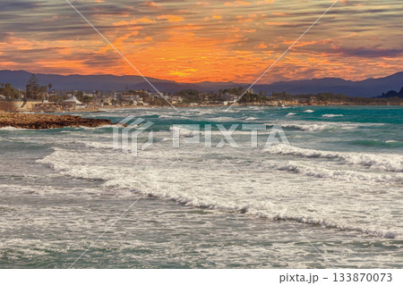 Orange sunset over sea waves and mountains with coastal resort buildings Orange sunset over sea waves and mountains with coastal resort buildings 133870073
