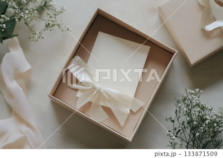 Minimalist flat lay of open gift box with blank greeting card, cream ribbon bow and baby's breath on neutral pastel background 133871509