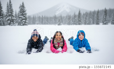 Three happy children of different ethnicities are playing in the snow, smiling and looking at the camera, enjoying outdoor winter activities in a snowy forest landscape with mountains 133872145