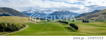 Stunning panorama of new zealand's countryside features lush green meadows, a winding road, rows of trees, and majestic snow capped mountains under a cloudy sky 133872313