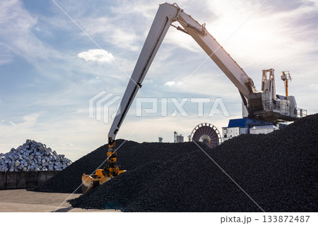 Large industrial crane loads dark coal piles bright sunny sky backlight background. Metallic blocks and massive equipment storage site. Energy commodity industry global trade logistics business Large industrial crane loads dark coal piles bright sunny sky backlight background. Metallic blocks and massive equipment storage site. Energy commodity industry global trade logistics business 133872487