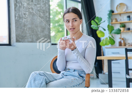 Teenage girl sitting at home in chair with smartphone, recording an audio message 133872858