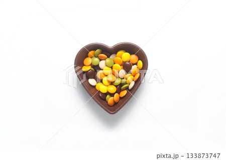 multi-colored sweet candies in heart shaped bowl on white background, top view. Space for text multi-colored sweet candies in heart shaped bowl on white background, top view. Space for text 133873747