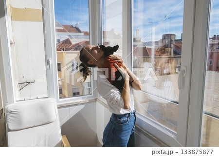 Joyful woman twirls with pet dog by large window viewing sky 133875877