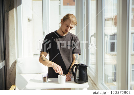 Male student taking short break with warm beverage by sunny window Male student taking short break with warm beverage by sunny window 133875902