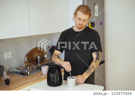Tattooed individual blends coffee using metal spoon in kitchen setting Tattooed individual blends coffee using metal spoon in kitchen setting 133875924