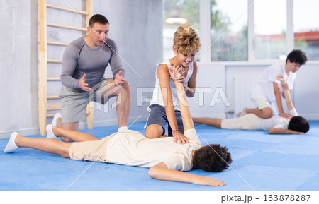 Young schoolboy practicing basic self-defense moves during training, practicing painful pronating wristlock on opponent lying face down on floor at gym Young schoolboy practicing basic self-defense moves during training, practicing painful pronating wristlock on opponent lying face down on floor at gym 133878287