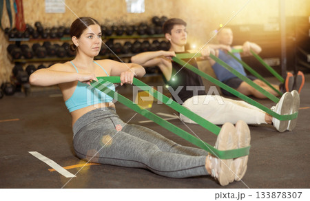 Motivated young woman practicing exercises with stretch rope sitting near other people in gym 133878307