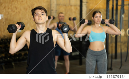 Young dedicated active strong fit man doing exercises for shoulders while pushing up dumbbells in CrossFit center Young dedicated active strong fit man doing exercises for shoulders while pushing up dumbbells in CrossFit center 133879184