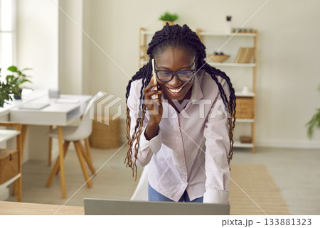 Happy young African American businesswoman using laptop and talking on mobile phone Happy young African American businesswoman using laptop and talking on mobile phone 133881323
