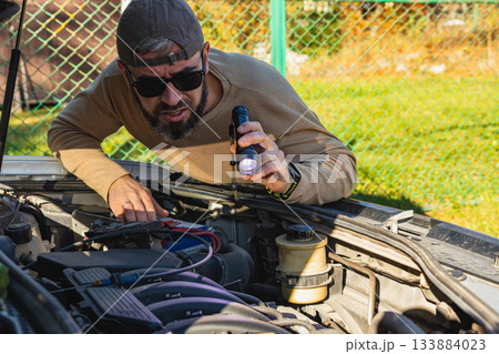 Man examines car engine in outdoor workshop with flashlight 133884023