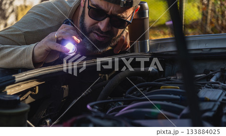 Man inspecting car engine with flashlight in outdoor setting during daylight 133884025