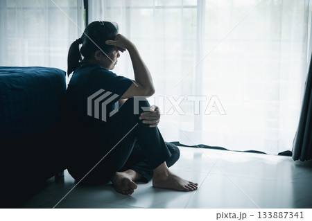 A lonely woman sits on the floor by window with hand on head in dark room. Concept of depression, sadness, anxiety, stress, mental health problem, loneliness and emotional struggle in life. 133887341