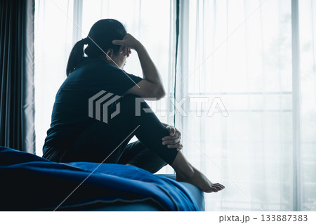 A stressed woman sitting alone on bed in dark room, holding her head down, symbolizing depression, anxiety, hopelessness, loneliness, mental health struggle, burnout and emotional pain. 133887383