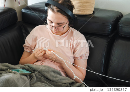 Teenager girl wearing glasses and headphones sitting comfortably at home, knitting using a colorful yarn, following a pattern or scheme displayed on her smartphone. Cozy creative hobby scene. 133890857