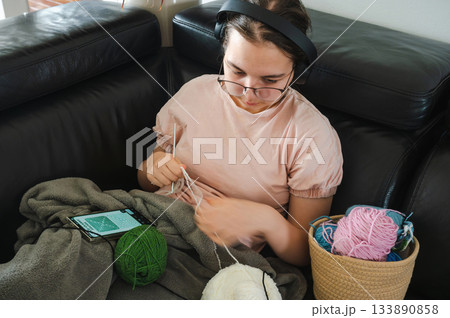Teenager girl wearing glasses and headphones sitting comfortably at home, knitting using a colorful yarn, following a pattern or scheme displayed on her smartphone. Cozy creative hobby scene. 133890858