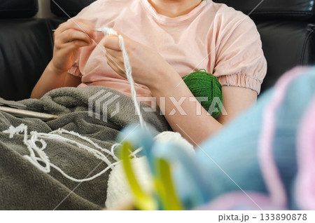 Teenager girl wearing glasses and headphones sitting comfortably at home, knitting using a colorful yarn, following a pattern or scheme displayed on her smartphone. Cozy creative hobby scene. Teenager girl wearing glasses and headphones sitting comfortably at home, knitting using a colorful yarn, following a pattern or scheme displayed on her smartphone. Cozy creative hobby scene. 133890878