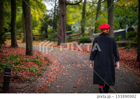 Elegant, well-groomed woman with fashionable style walking in an autumn park on a cloudy day. She wears a red beret, red lipstick, and matching red tights, looking confident and sophisticated. 133890982