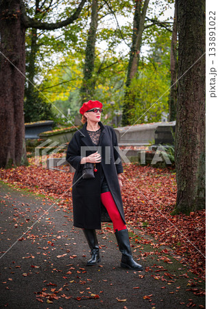 Elegant, well-groomed woman with fashionable style walking in an autumn park on a cloudy day. She wears a red beret, red lipstick, and matching red tights, looking confident and sophisticated. Elegant, well-groomed woman with fashionable style walking in an autumn park on a cloudy day. She wears a red beret, red lipstick, and matching red tights, looking confident and sophisticated. 133891022