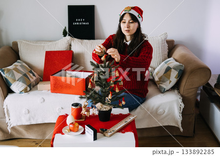 A young woman in a Santa hat and red sweater sits on a sofa decorating a small tabletop Christmas tree. Festive decor, small space living, generational traditions, seasonal homemaking. A young woman in a Santa hat and red sweater sits on a sofa decorating a small tabletop Christmas tree. Festive decor, small space living, generational traditions, seasonal homemaking. 133892285