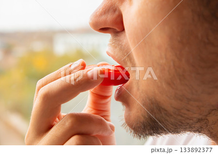 A young man holds a red tomato slice close to his mouth, preparing to take a bite while sitting indoors by a window. Plum tomato A young man holds a red tomato slice close to his mouth, preparing to take a bite while sitting indoors by a window. Plum tomato 133892377