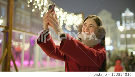 A young beautiful girl in a red coat walks in the evening at the European Christmas Market and talks on the phone against the backdrop of an ice rink with illumination and garlands. A young beautiful girl in a red coat walks in the evening at the European Christmas Market and talks on the phone against the backdrop of an ice rink with illumination and garlands. 133894036
