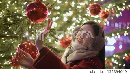 A young beautiful girl in a red coat stands near a Christmas tree with garlands and illuminations in the evening at the European Christmas Market and touches the Christmas tree decorations. 133894049