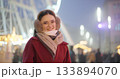 A young beautiful girl in a red coat walks in the evening at the New Year's Christmas fair against the backdrop of a Ferris wheel among lights and garlands and admires the decorations 133894070