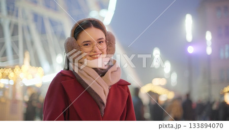 A young beautiful girl in a red coat walks in the evening at the New Year's Christmas fair against the backdrop of a Ferris wheel among lights and garlands and admires the decorations 133894070