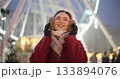 A young beautiful girl in a red coat walks in the evening at the New Year's Christmas fair against the backdrop of a Ferris wheel among lights and garlands and admires the decorations 133894076