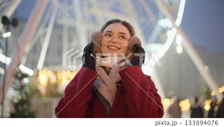 A young beautiful girl in a red coat walks in the evening at the New Year's Christmas fair against the backdrop of a Ferris wheel among lights and garlands and admires the decorations A young beautiful girl in a red coat walks in the evening at the New Year's Christmas fair against the backdrop of a Ferris wheel among lights and garlands and admires the decorations 133894076