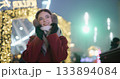 A young beautiful girl in a red coat walks in the evening at the New Year's Christmas fair against the backdrop of a Ferris wheel among lights and garlands and admires the decorations 133894084