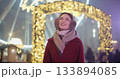 A young beautiful girl in a red coat walks in the evening at the New Year's Christmas fair against the backdrop of a Ferris wheel among lights and garlands and admires the decorations 133894085