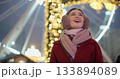 A young beautiful girl in a red coat walks in the evening at the New Year's Christmas fair against the backdrop of a Ferris wheel among lights and garlands and admires the decorations 133894089